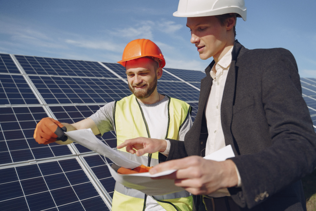 Technicien en gilet jaune et casque orange présentant un plan à un client lors d'un audit énergétique devant des panneaux solaires