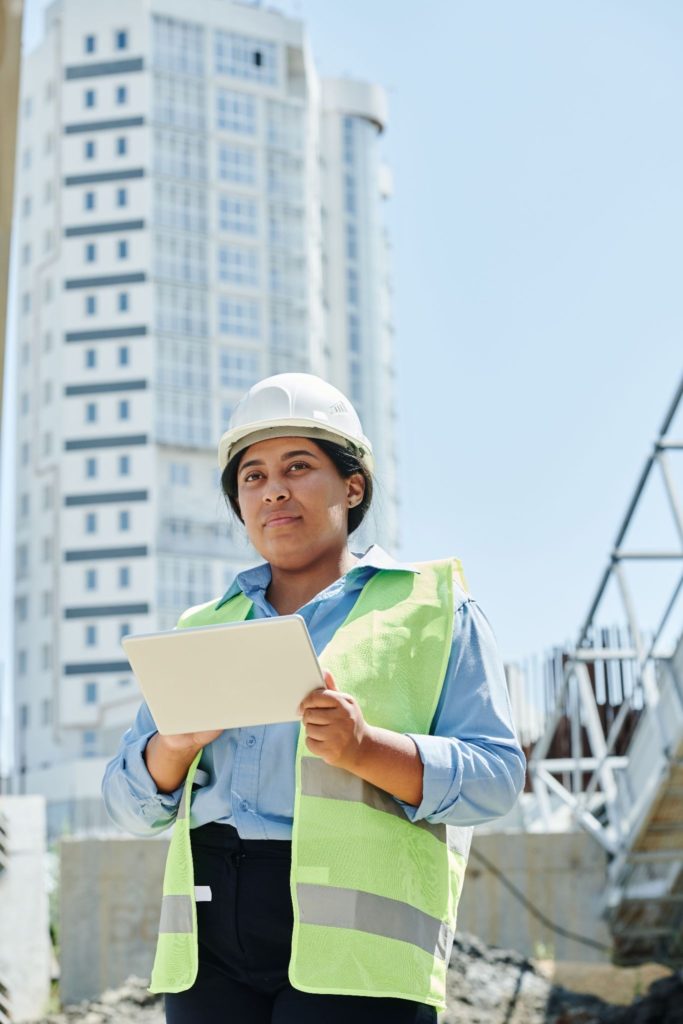 Technicienne en gilet jaune et casque blanc tenant une tablette lors d'un audit énergétique devant un immeuble résidentiel